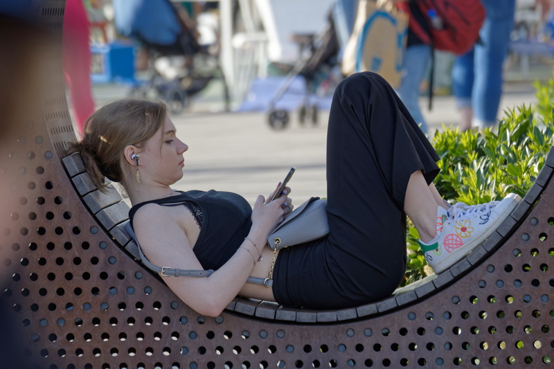 6 - A girl wearing a black sleeveless top, wide-leg trousers, and patterned sneakers while relaxing with a smartphone on an urban curved bench